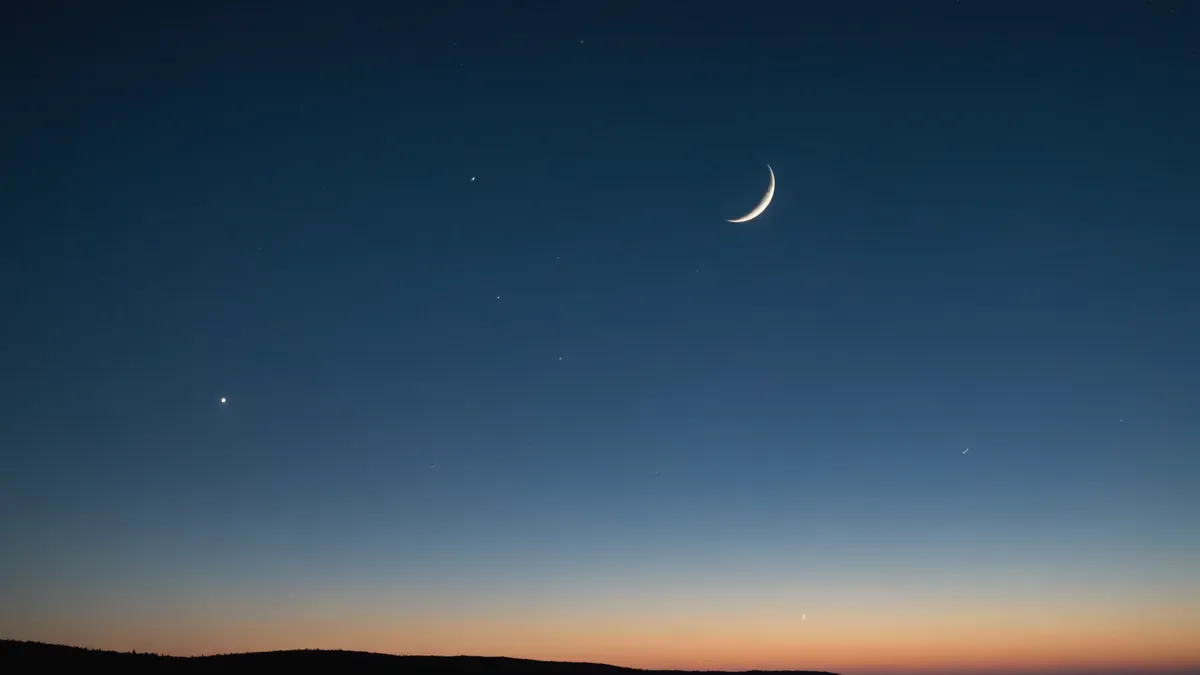 Fin croissant de Lune dans le ciel bleu profond du crépuscule, au-dessus d'une ligne de collines sombres et d'un horizon orangé